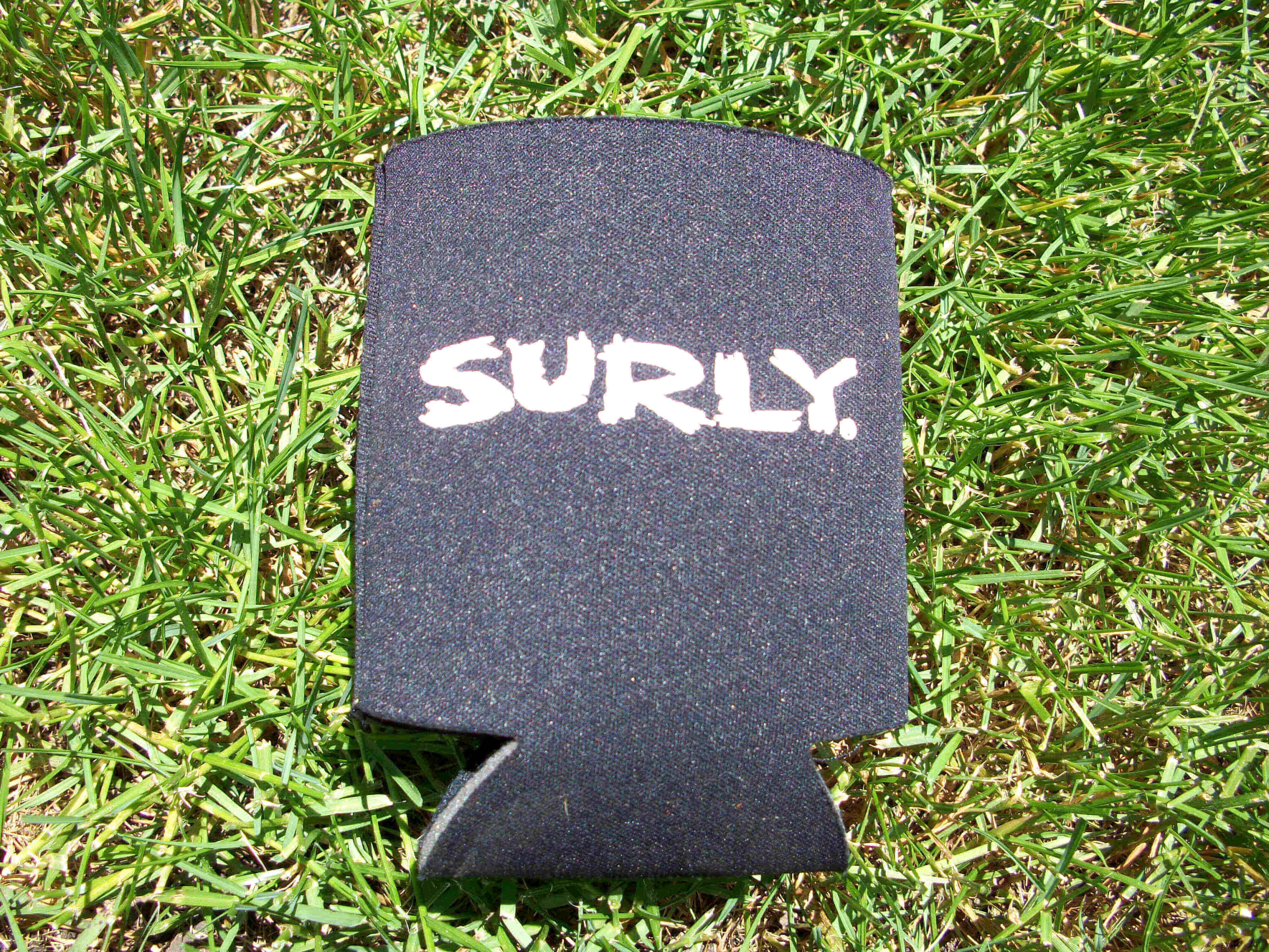 Downward view of a flattened, black Surly can cooler with a white logo graphic, laying on grass